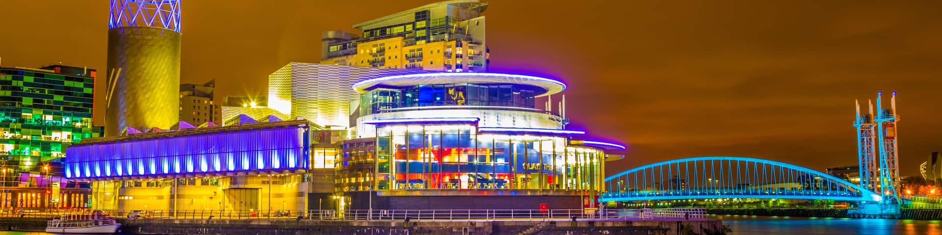 A brightly lit waterfront with modern buildings, a distinctive tower, and a blue-lit bridge.