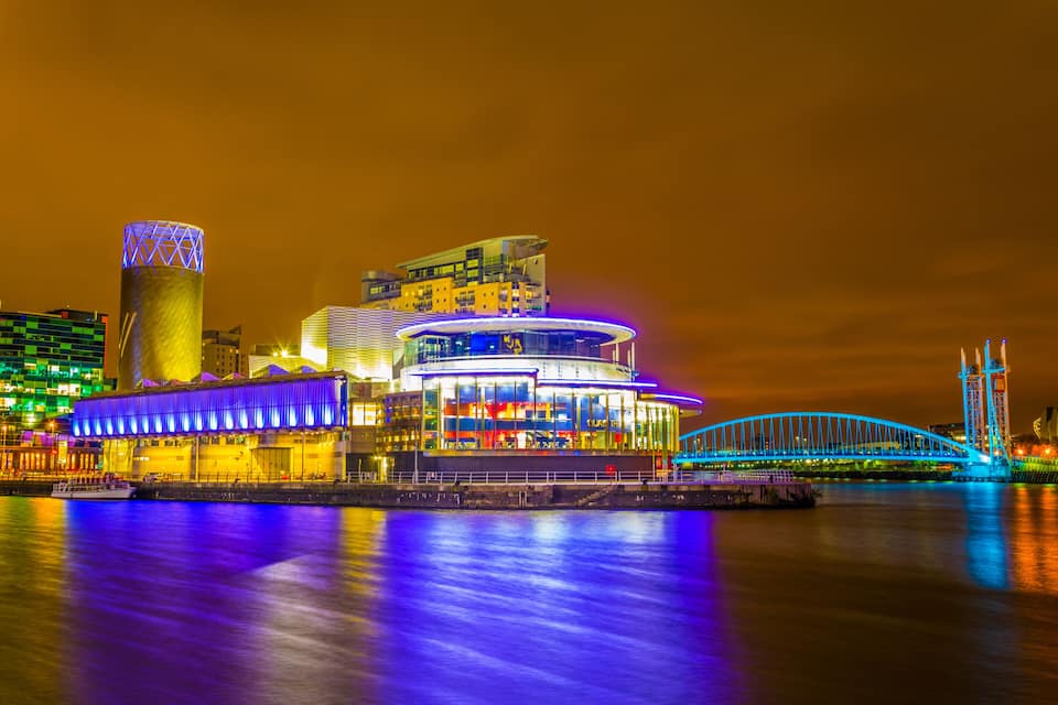 A brightly lit waterfront with modern buildings, a distinctive tower, and a blue-lit bridge.