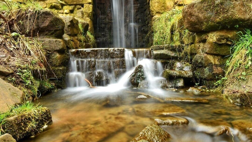 A stone bridge over a flowing stream with a waterfall in the background.
