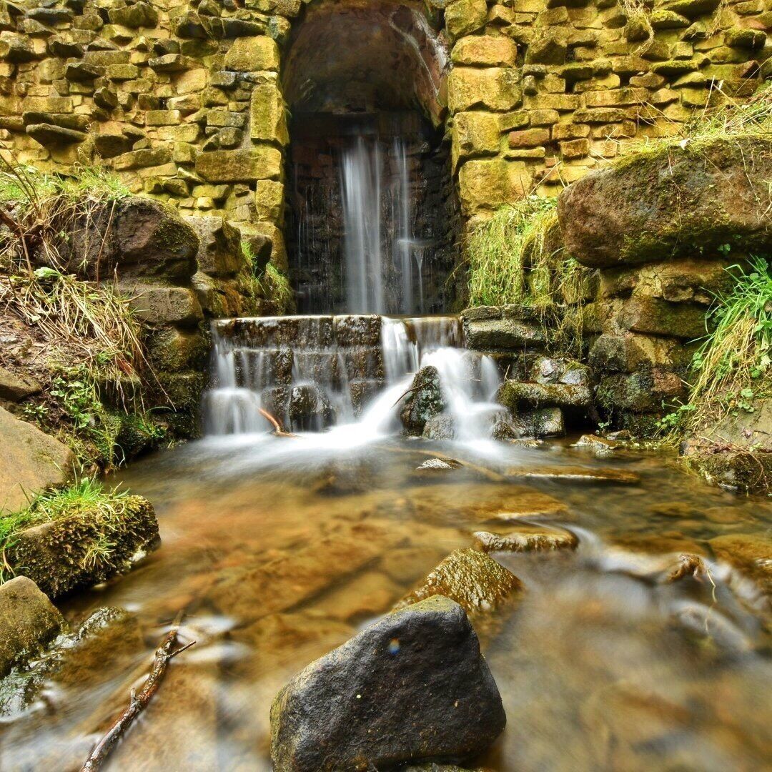 A stone bridge over a flowing stream with a waterfall in the background.