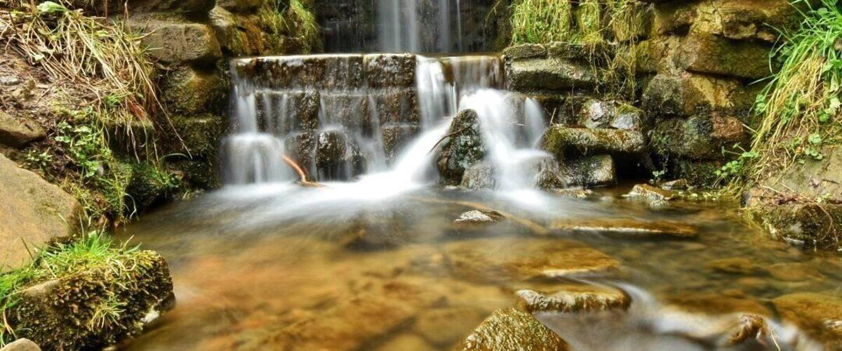 A stone bridge over a flowing stream with a waterfall in the background.
