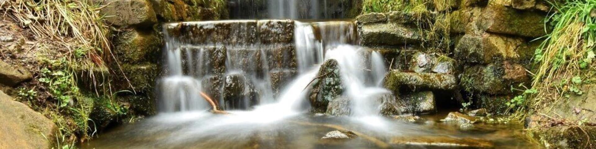 A stone bridge over a flowing stream with a waterfall in the background.