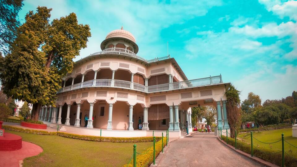 A large, ornate building with a dome and multiple balconies, surrounded by well-maintained gardens and a clear sky.