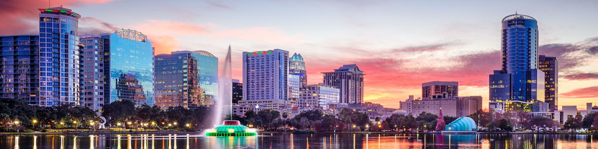 A city skyline at sunset with illuminated buildings and a calm lake reflecting the scene.