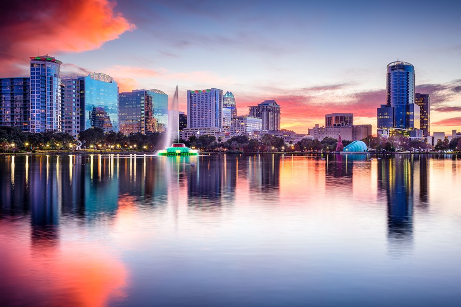 A city skyline at sunset with illuminated buildings and a calm lake reflecting the scene.