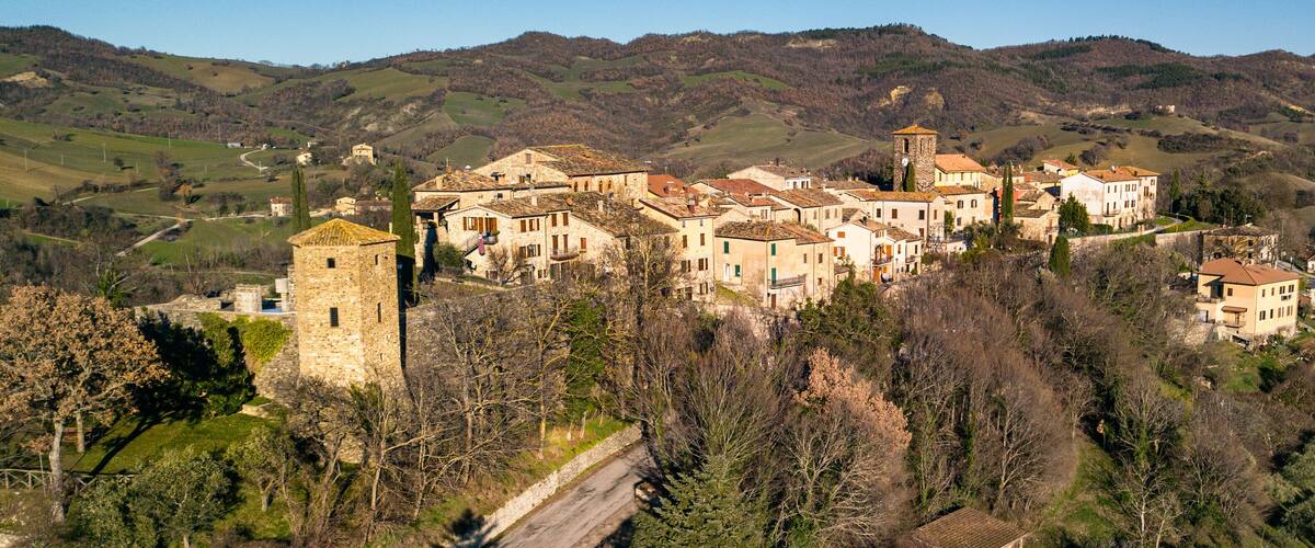A historic village with stone buildings and a prominent tower, surrounded by rolling hills and sparse trees.