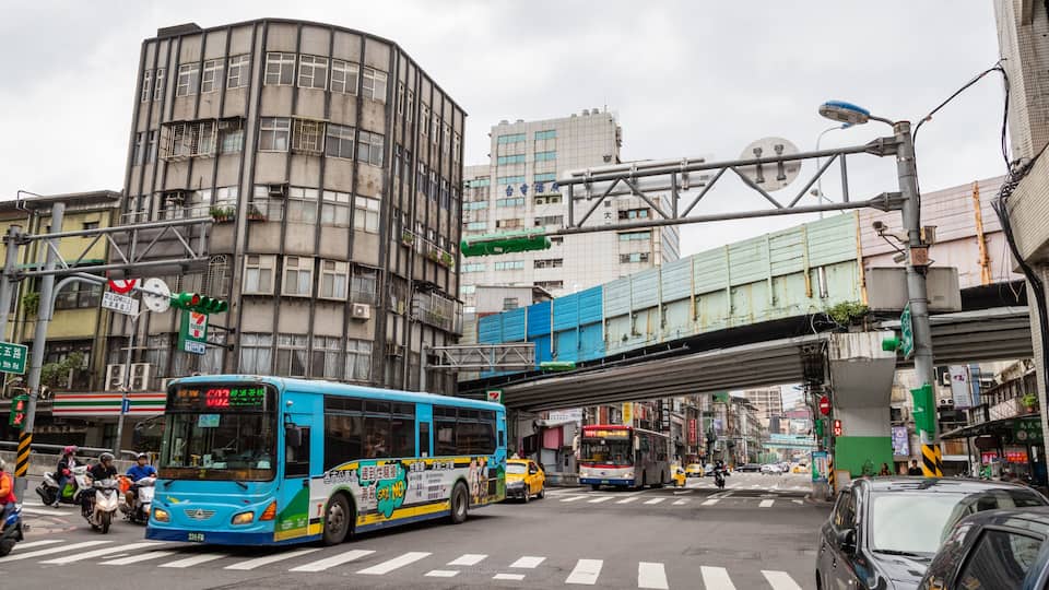 A busy urban street with a blue bus, a pedestrian overpass, and various buildings.