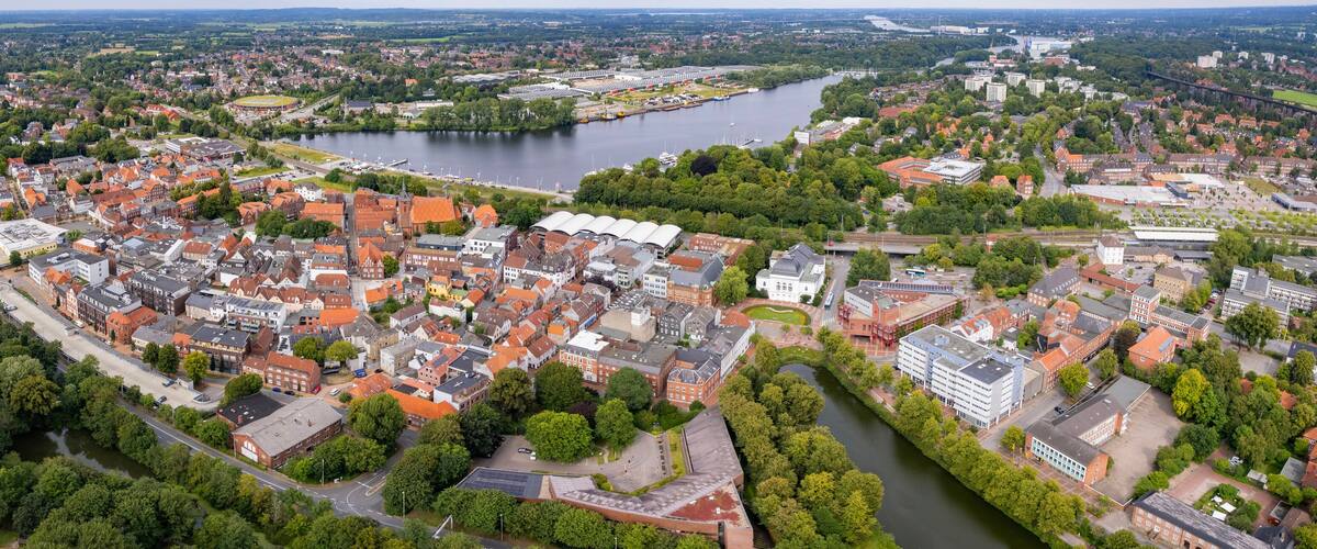An aerial view of a town with a river, surrounded by greenery and red-roofed buildings.