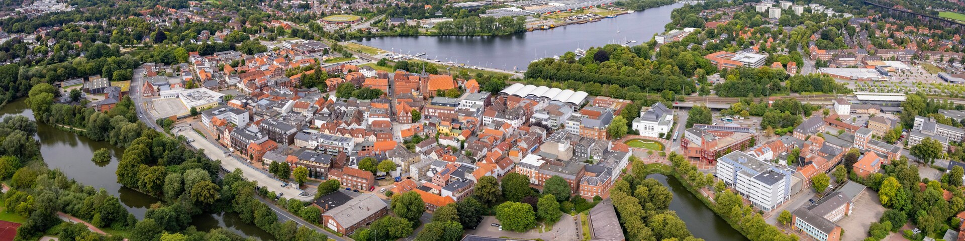 An aerial view of a town with a river, surrounded by greenery and red-roofed buildings.