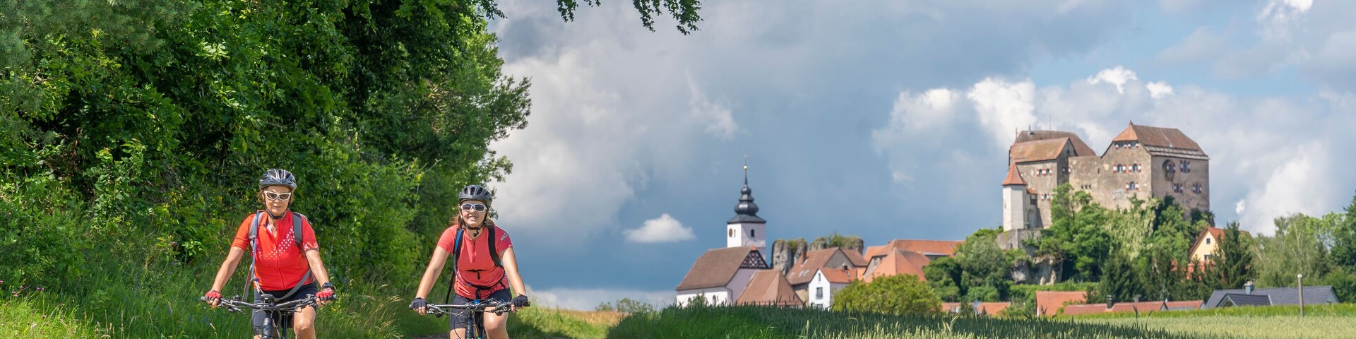 Two cyclists on a dirt path with a church and castle in the background.