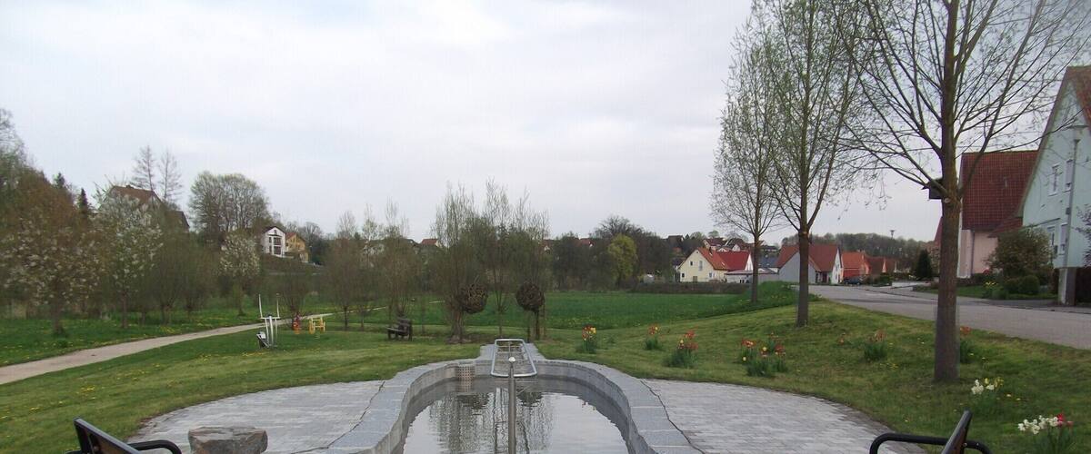 A circular water feature with benches, surrounded by a paved area and a grassy field with trees and flowers.