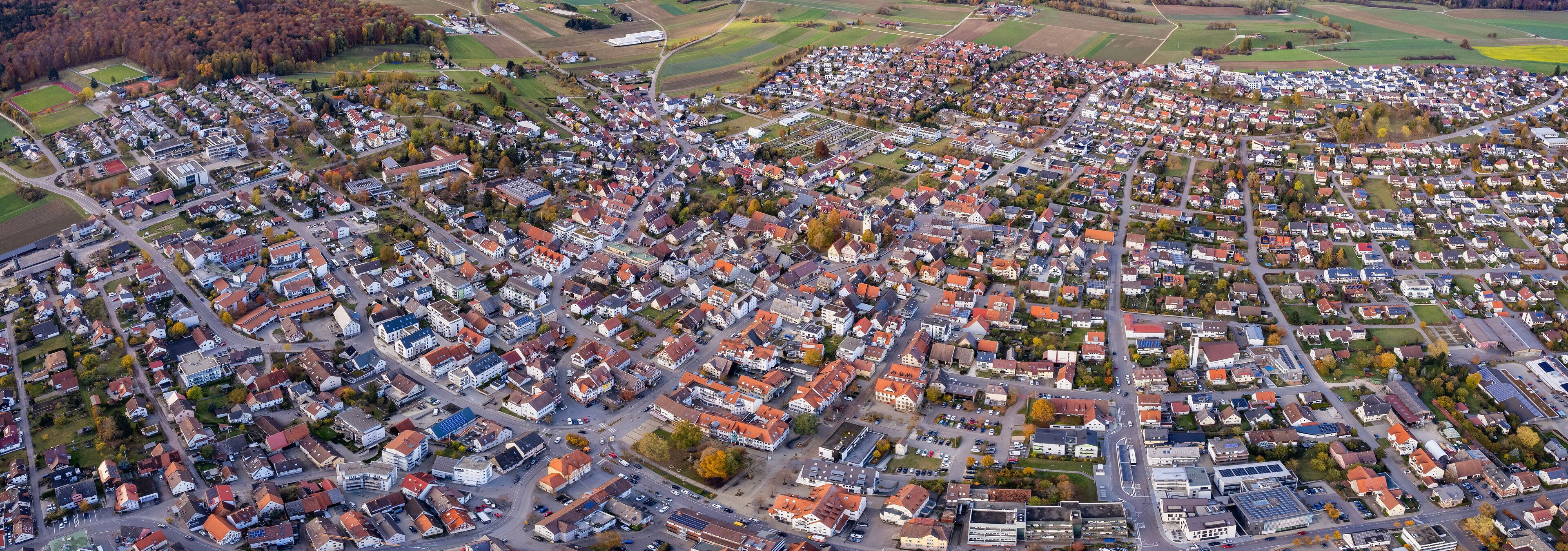 An aerial view of a densely populated residential area with a mix of houses and small buildings, surrounded by fields and forests.