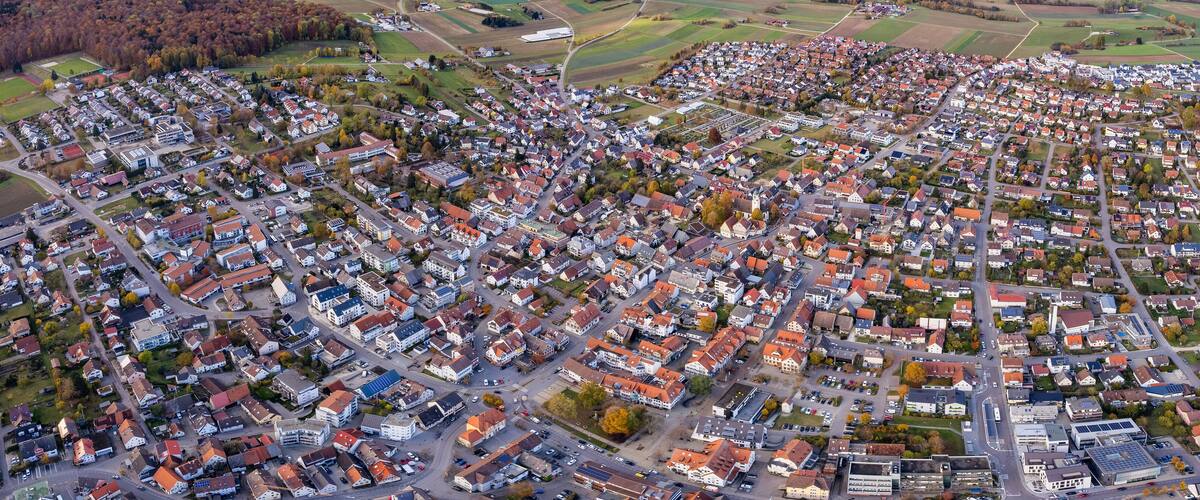 An aerial view of a densely populated residential area with a mix of houses and small buildings, surrounded by fields and forests.