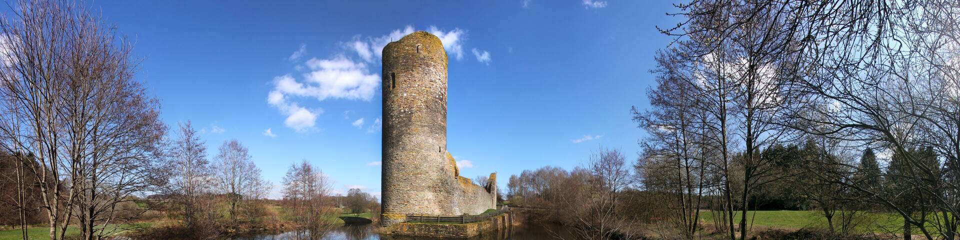 A stone tower by a calm river, surrounded by trees and a grassy field.
