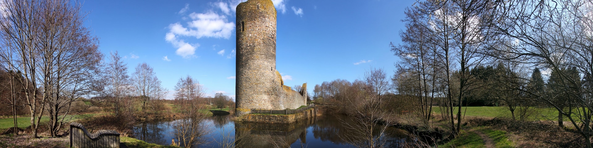 Una torre de piedra junto a un río tranquilo, rodeada de árboles y un campo cubierto de hierba.