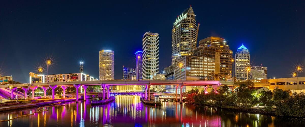 A nighttime cityscape with illuminated skyscrapers and a bridge reflecting on the water.