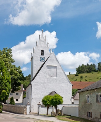 A white church with a clock tower and a cross on top, surrounded by trees and houses.