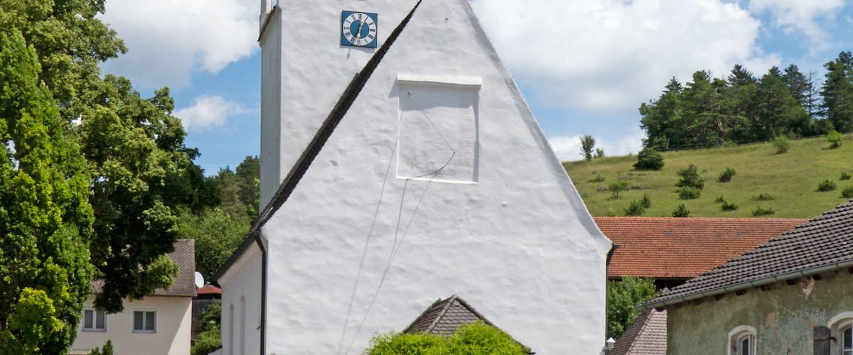 A white church with a clock tower and a cross on top, surrounded by trees and houses.