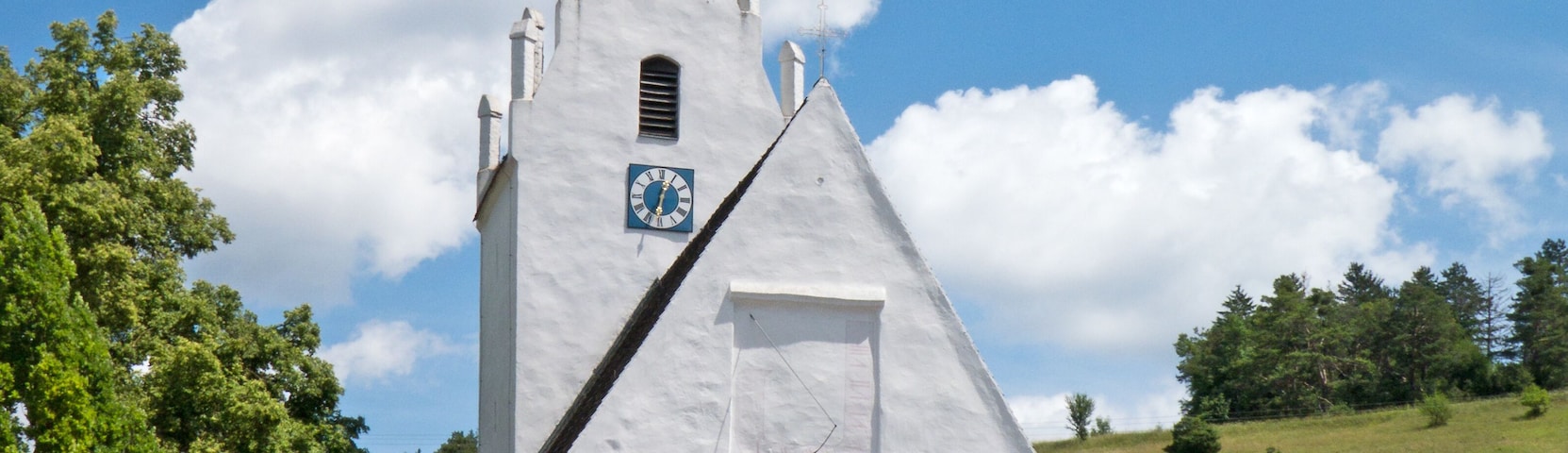 A white church with a clock tower and a cross on top, surrounded by trees and houses.