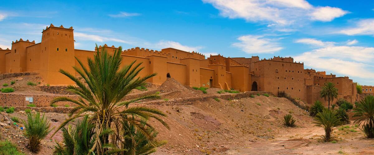 A large fortress with multiple towers and walls, surrounded by palm trees and rocky terrain.