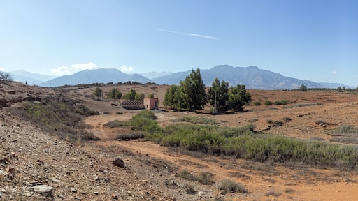 Moroccan countryside scene: red earth, sparse vegetation, a pink house, and distant mountains under a serene sky.