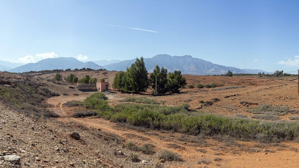 Moroccan countryside scene: red earth, sparse vegetation, a pink house, and distant mountains under a serene sky.