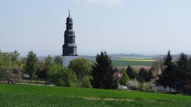 A church tower with a spire, surrounded by green fields and trees.