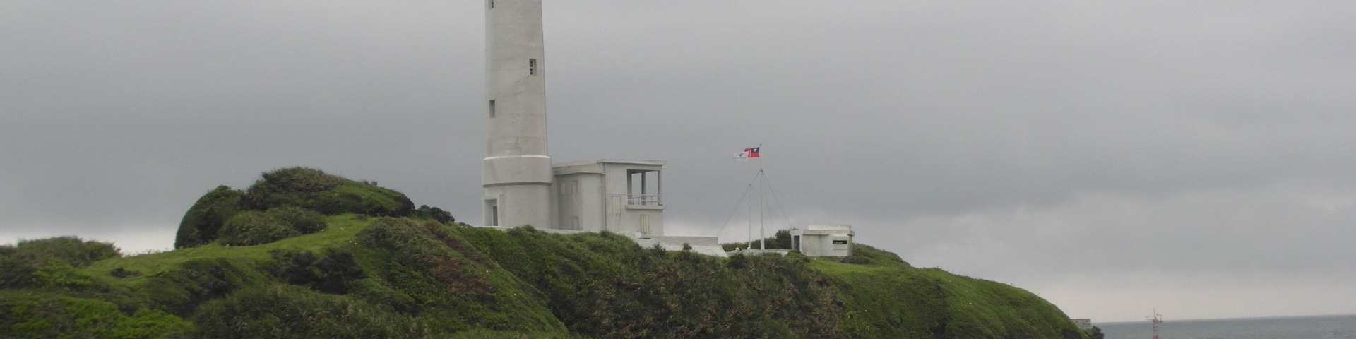 A white lighthouse on a grassy hill with a flag.