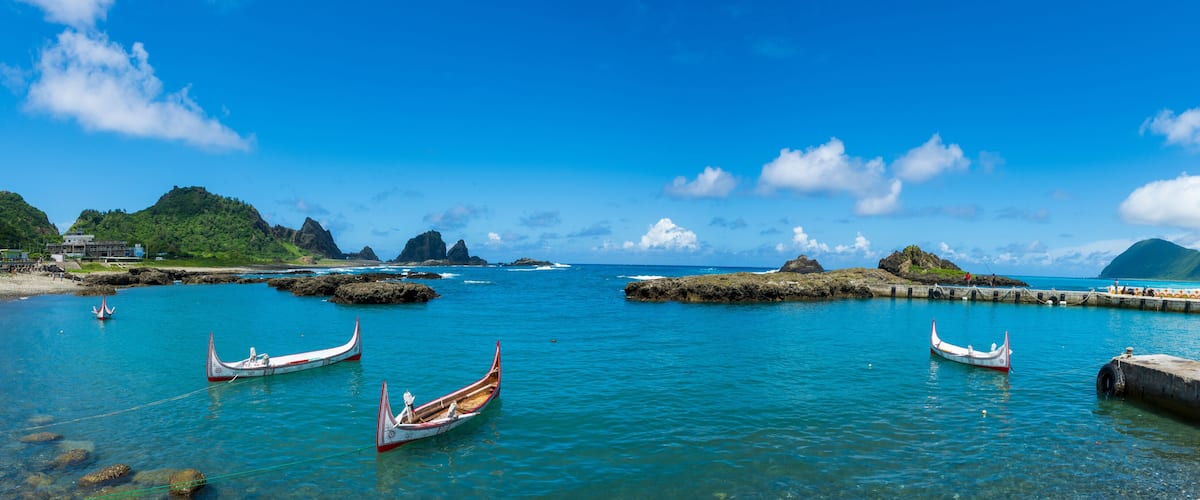 A coastal scene with clear turquoise waters, several traditional boats, rocky outcrops, and a pier with a railing.