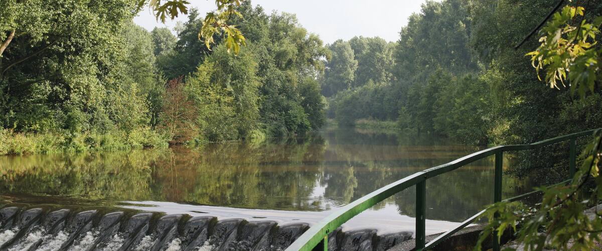 A water flow structure with a green railing, surrounded by dense forest and a calm lake.