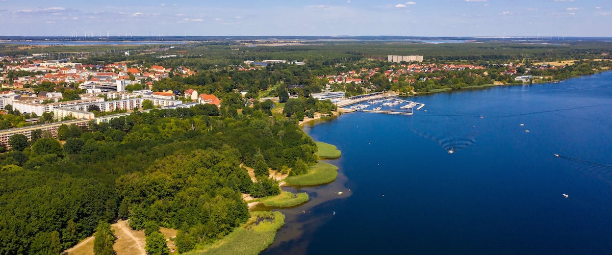 A coastal town with a marina, dense greenery, and wind turbines in the distance.