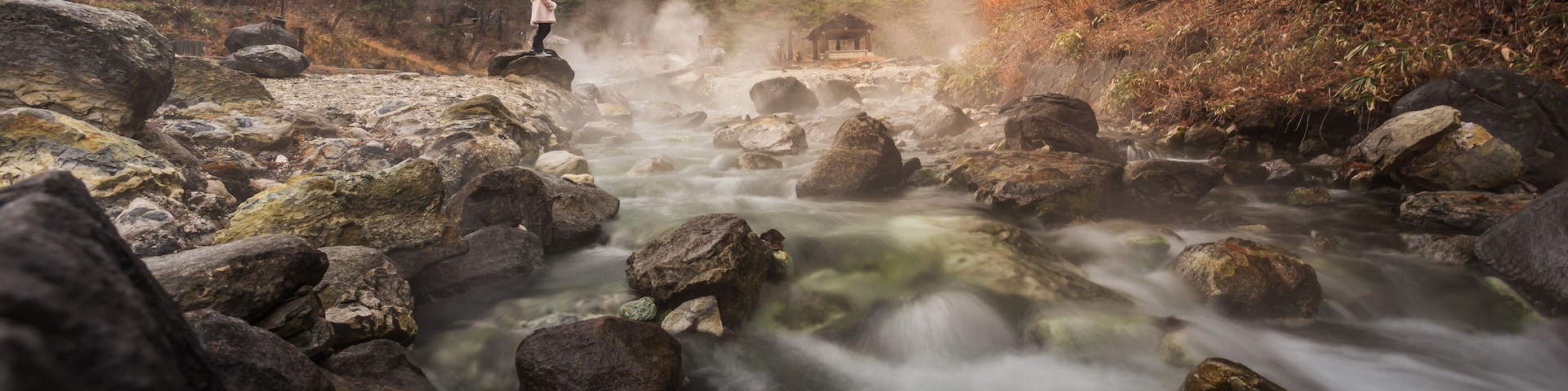 A rocky river with a person standing on a rock, surrounded by trees and a small building in the background.