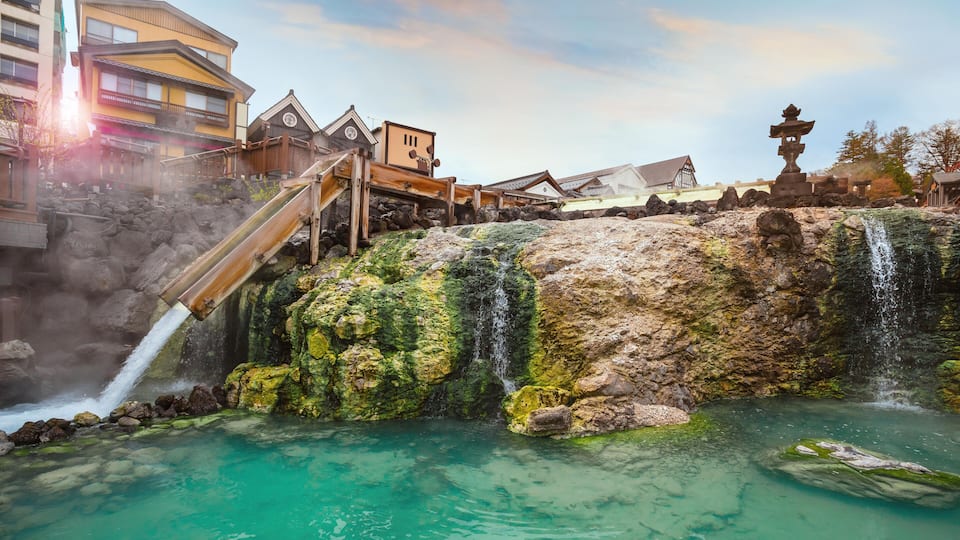 A traditional Japanese town with a waterfall, clear turquoise water, and wooden buildings.