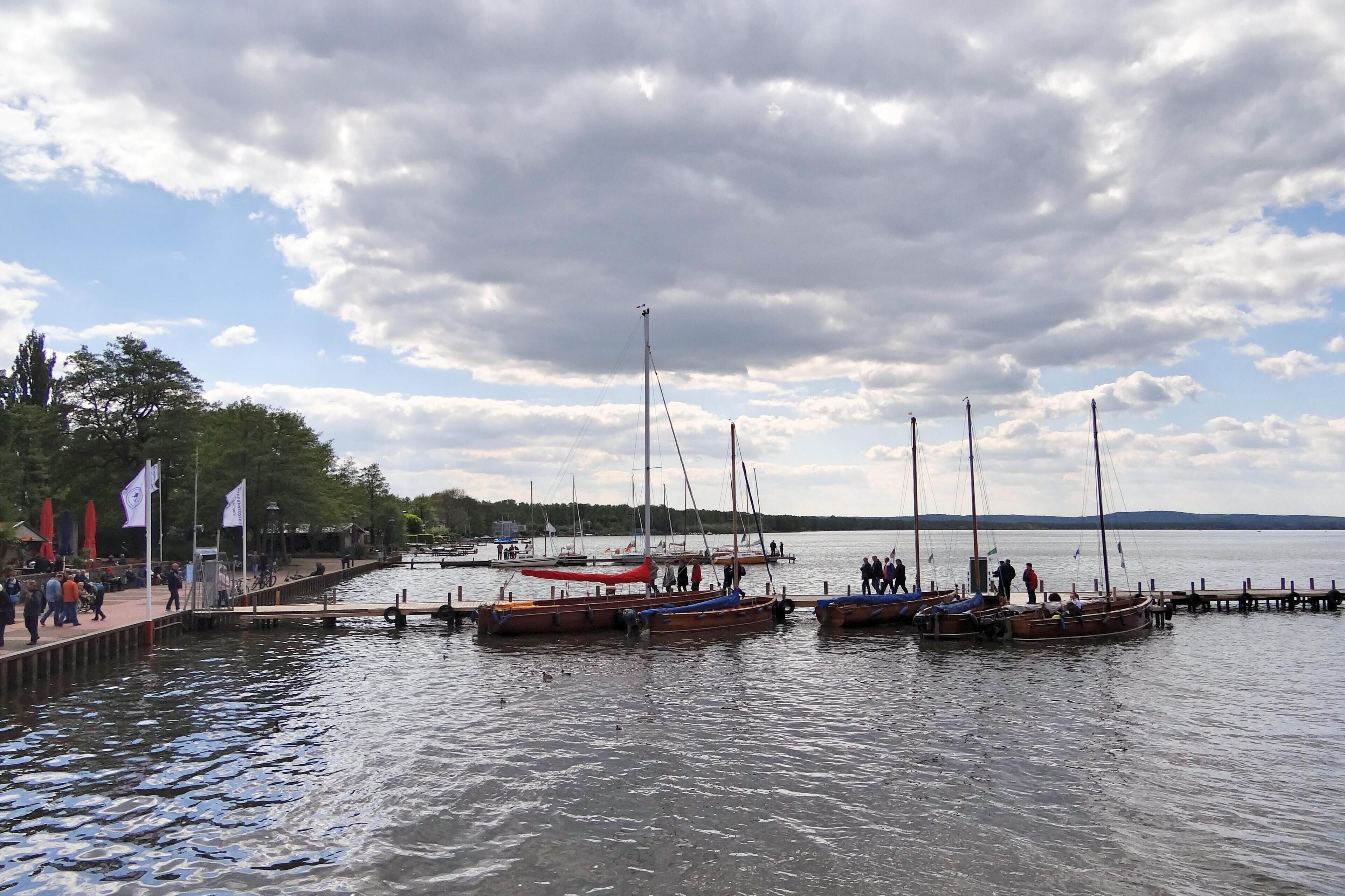 A lakeside pier with several boats docked, people walking, and a cloudy sky.