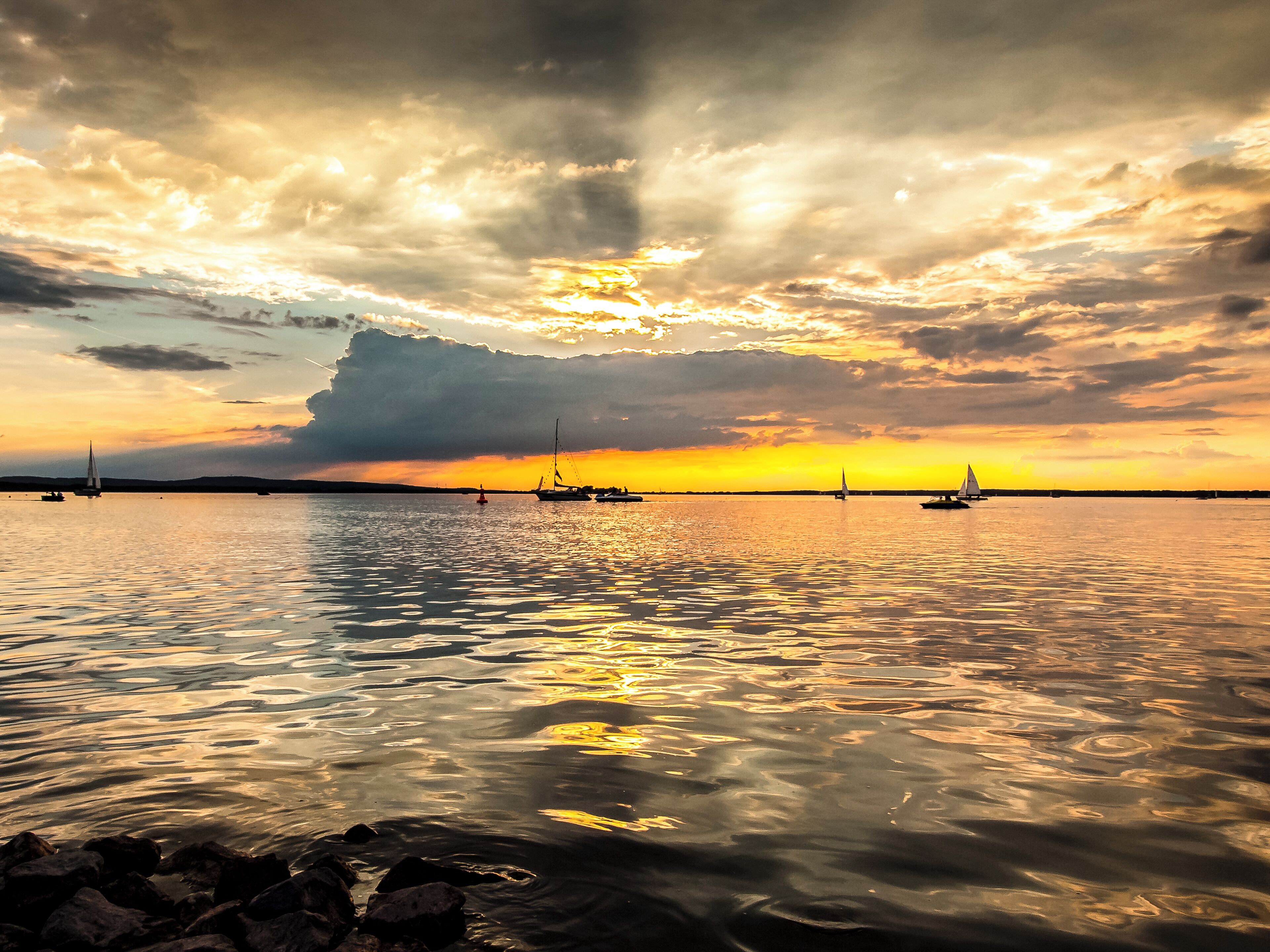 A sunset over a calm sea with sailboats and a large cloud formation.