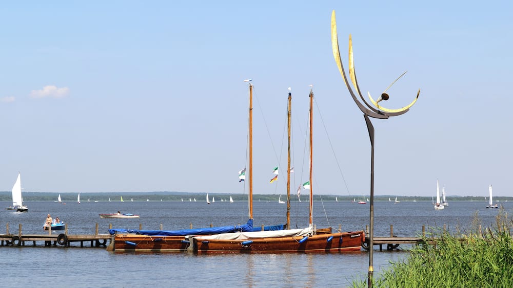 A calm lake with several boats docked at the pier, surrounded by greenery and sailboats in the distance.