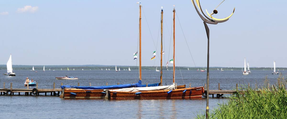 Un lago tranquilo con varios barcos amarrados en el muelle, rodeado de vegetación y veleros a lo lejos.