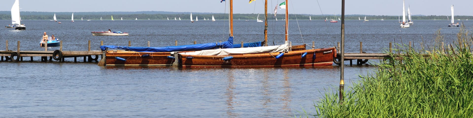 A calm lake with several boats docked at the pier, surrounded by greenery and sailboats in the distance.