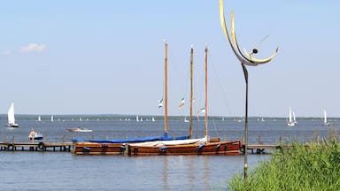 Um lago calmo com vários barcos atracados no cais, cercado por vegetação e veleiros ao longe.