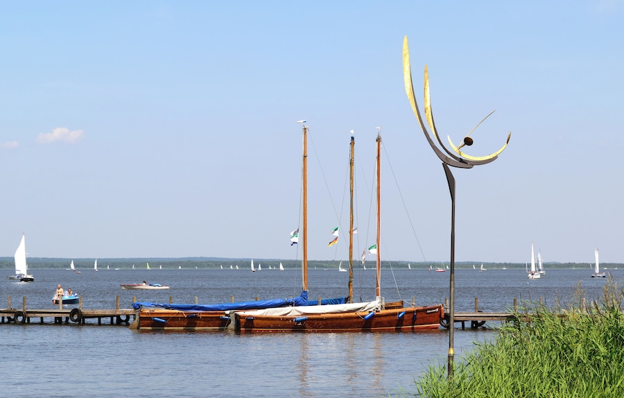 Un lago tranquilo con varios barcos amarrados en el muelle, rodeado de vegetación y veleros a lo lejos.