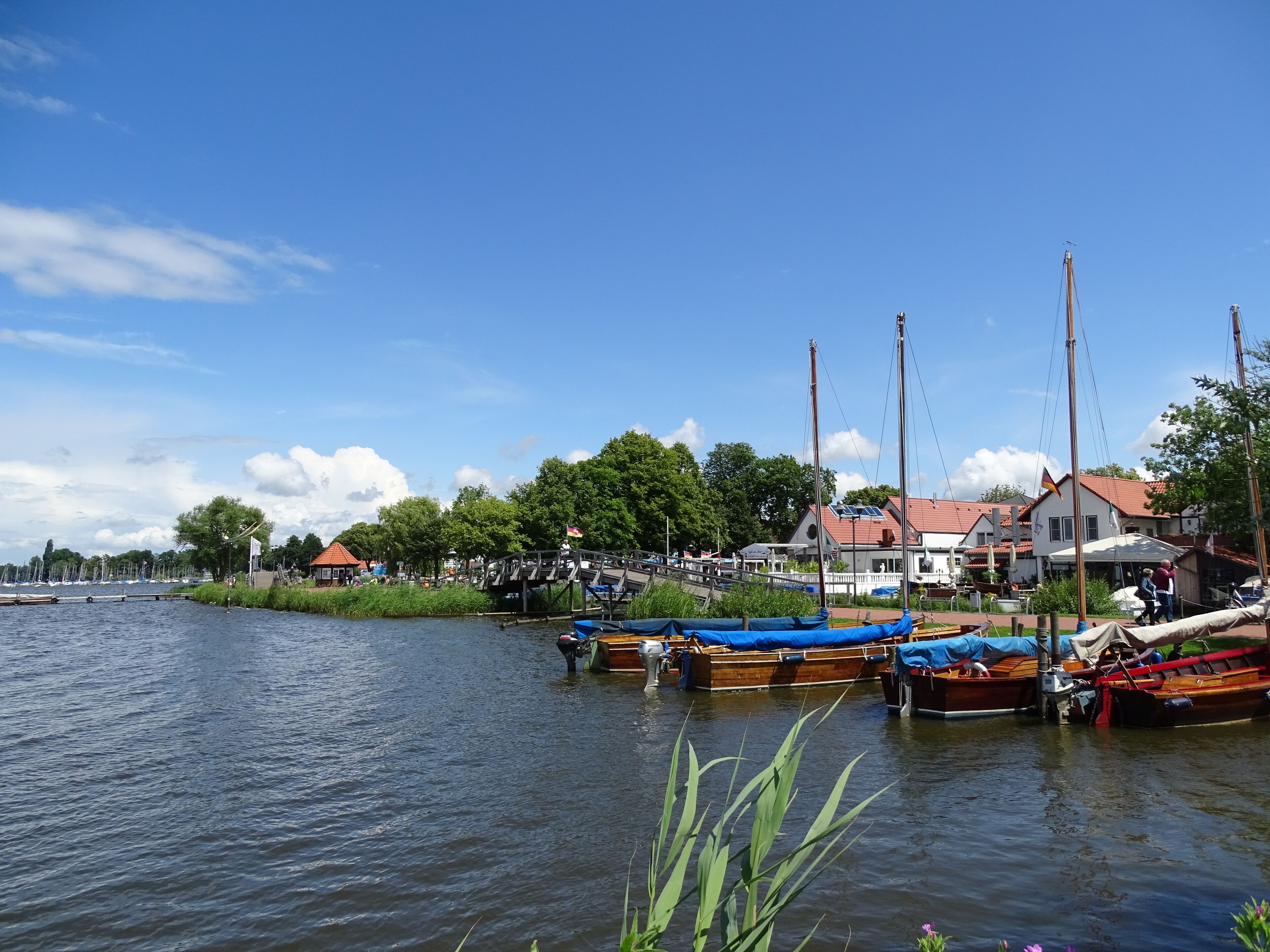 A calm river with boats docked along the shore, surrounded by trees and buildings.