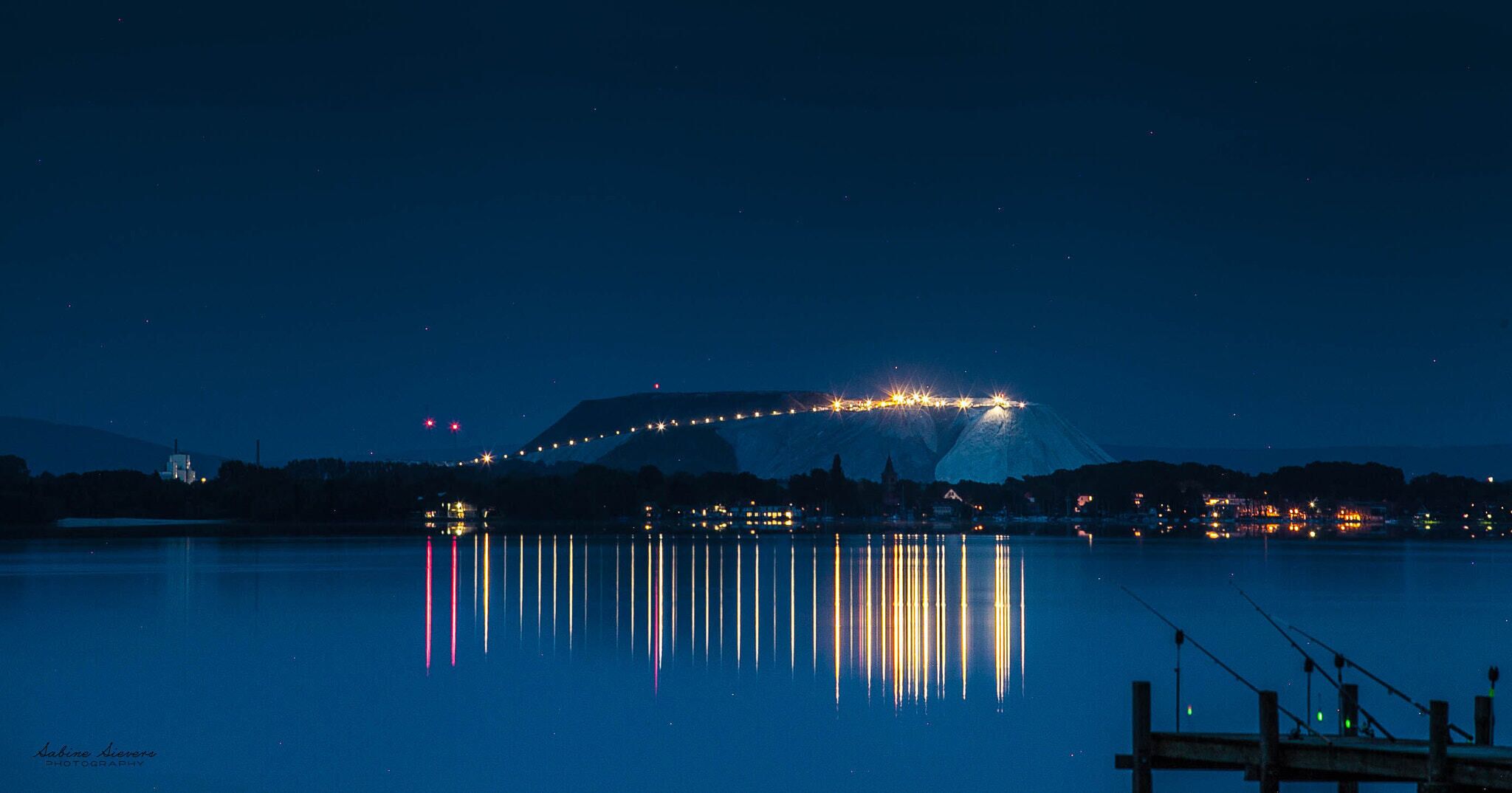 A nighttime view of a city with a mountain in the background and a bridge with lights reflecting on the water.