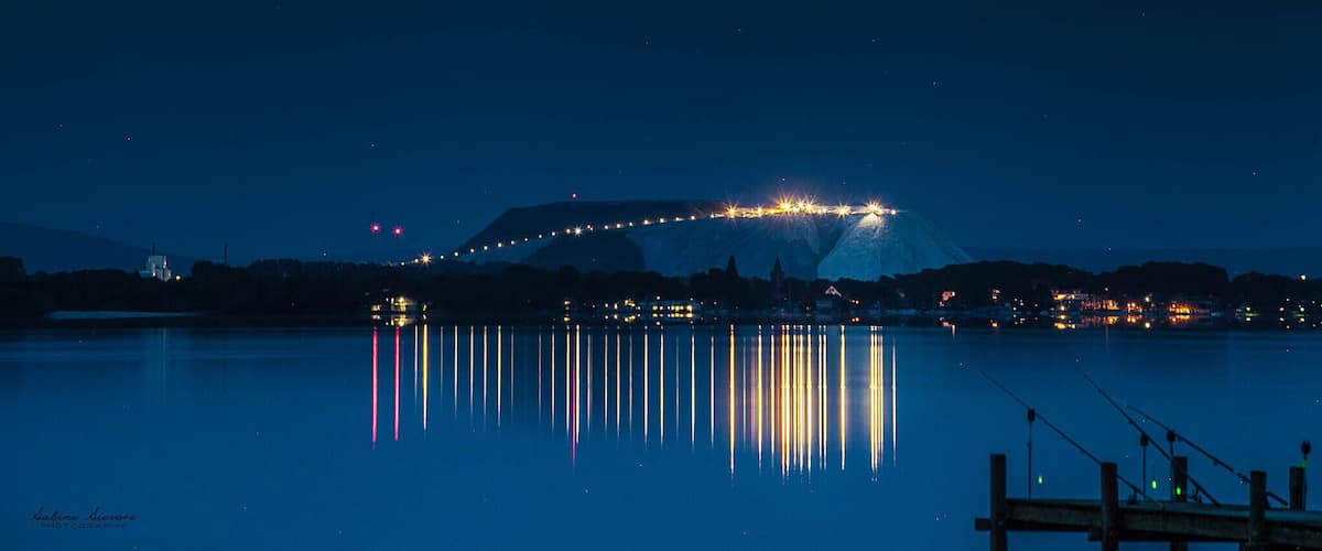 A nighttime view of a city with a mountain in the background and a bridge with lights reflecting on the water.