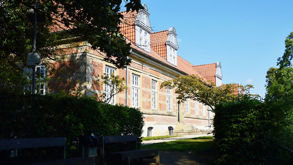 A historic building with red-tiled roofs, surrounded by lush greenery and a paved pathway.