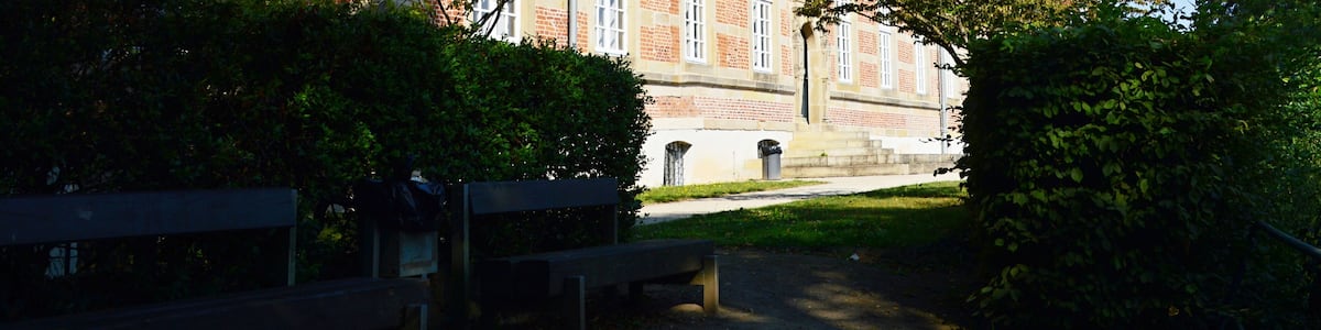 Un bâtiment historique, avec des toits recouverts de tuiles rouges, entouré d’une végétation luxuriante et bordé d’un chemin pavé.