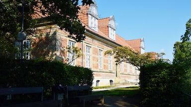 Um edifício histórico com telhados de telhas vermelhas, rodeado por vegetação exuberante e um caminho pavimentado.