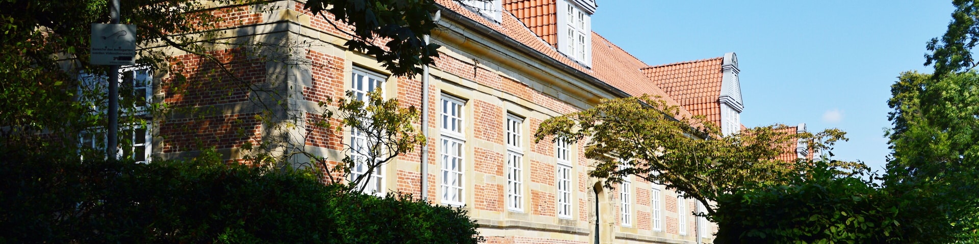Um edifício histórico com telhados de telhas vermelhas, rodeado por vegetação exuberante e um caminho pavimentado.
