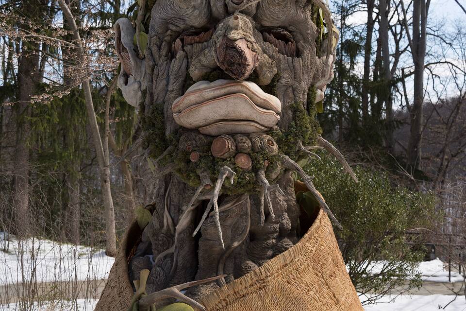 Una scultura di legno raffigurante un albero, con un grande frutto arancione e un sacco di iuta.