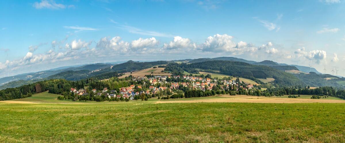 A village nestled in a valley with rolling hills and wind turbines in the distance.