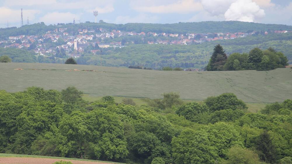 Eine hügelige Landschaft mit einem Dorf im Hintergrund, umgeben von dichten Wäldern und Feldern.